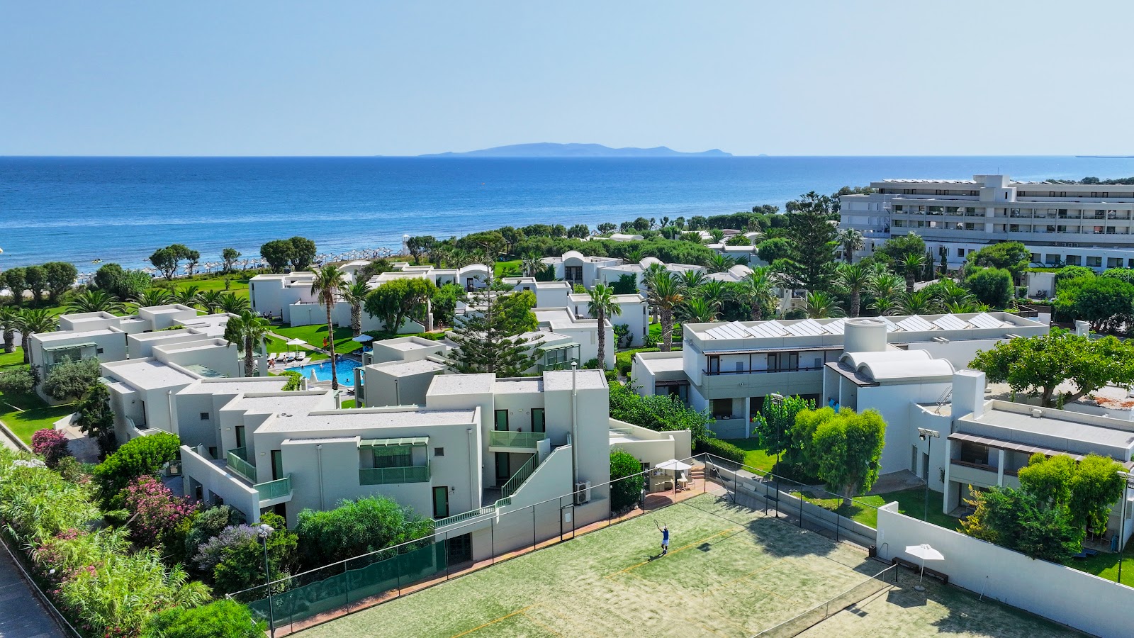 White villa-style buildings with flat roofs scattered across manicured grounds at Agapi Beach Resort, overlooking blue Mediterranean Sea and a distant island.
