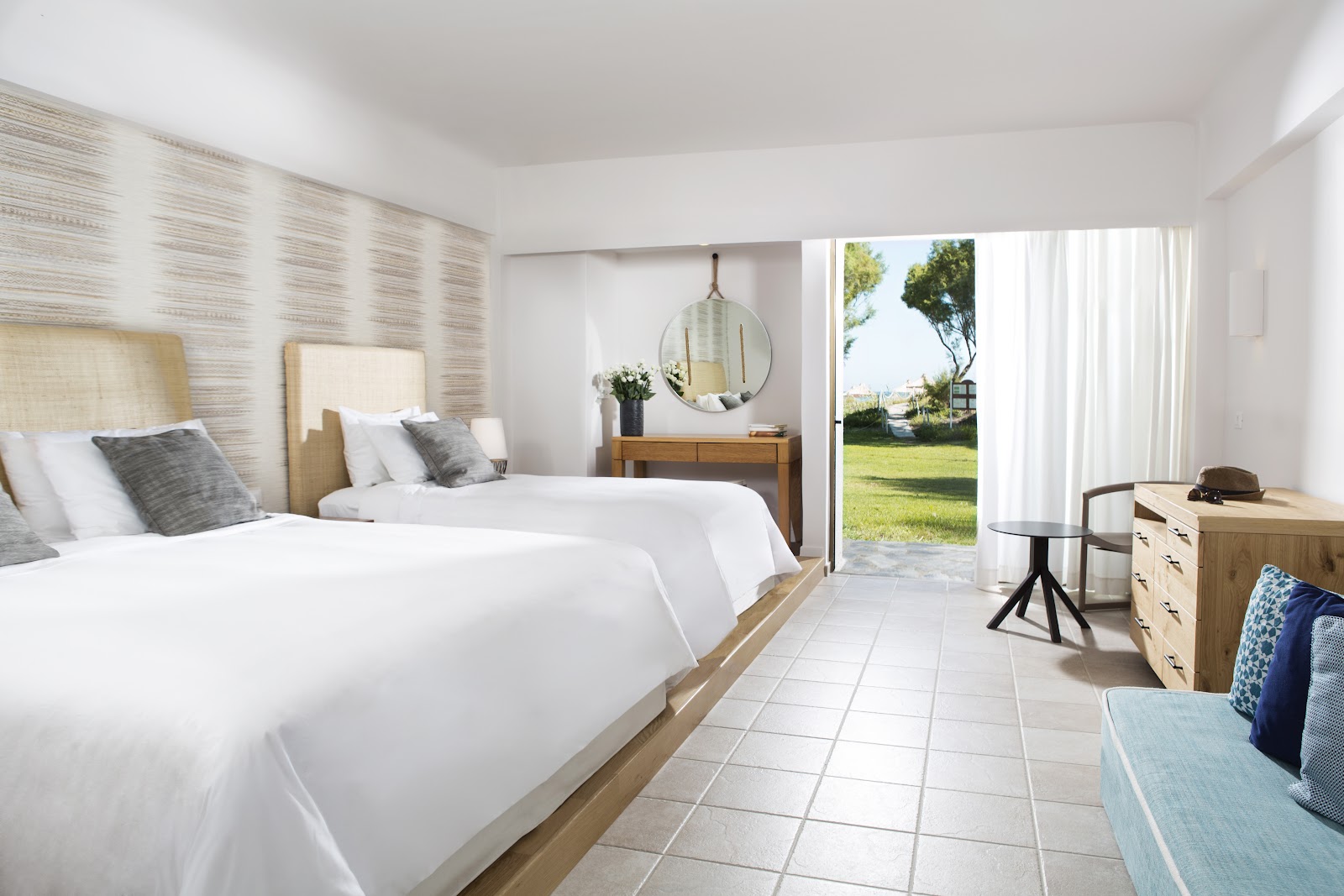 Modern resort bedroom with two queen beds, neutral furnishings, and sliding glass doors opening to a manicured lawn and trees.