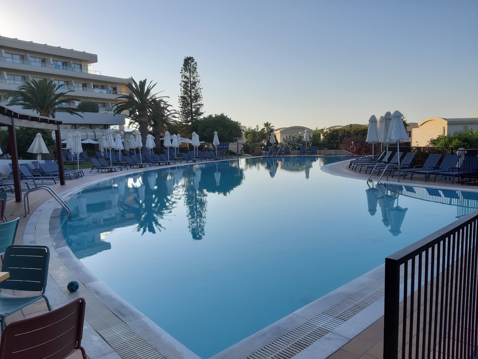 Large resort pool with white umbrellas and loungers, palm trees, and multi-story hotel building at Agapi Beach Resort under clear sky.