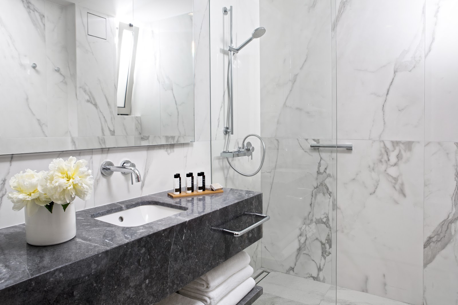 Modern bathroom at Agapi Beach Resort with marble walls, dark granite vanity, white flowers, and glass-enclosed shower with chrome fixtures.