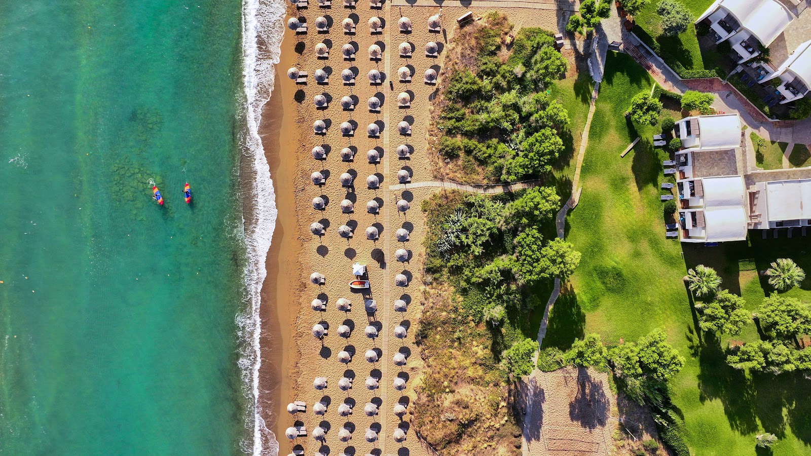 Aerial view of Agapi Beach Resort's sandy beach lined with umbrellas and loungers, turquoise water, and resort buildings nestled among green landscaping.