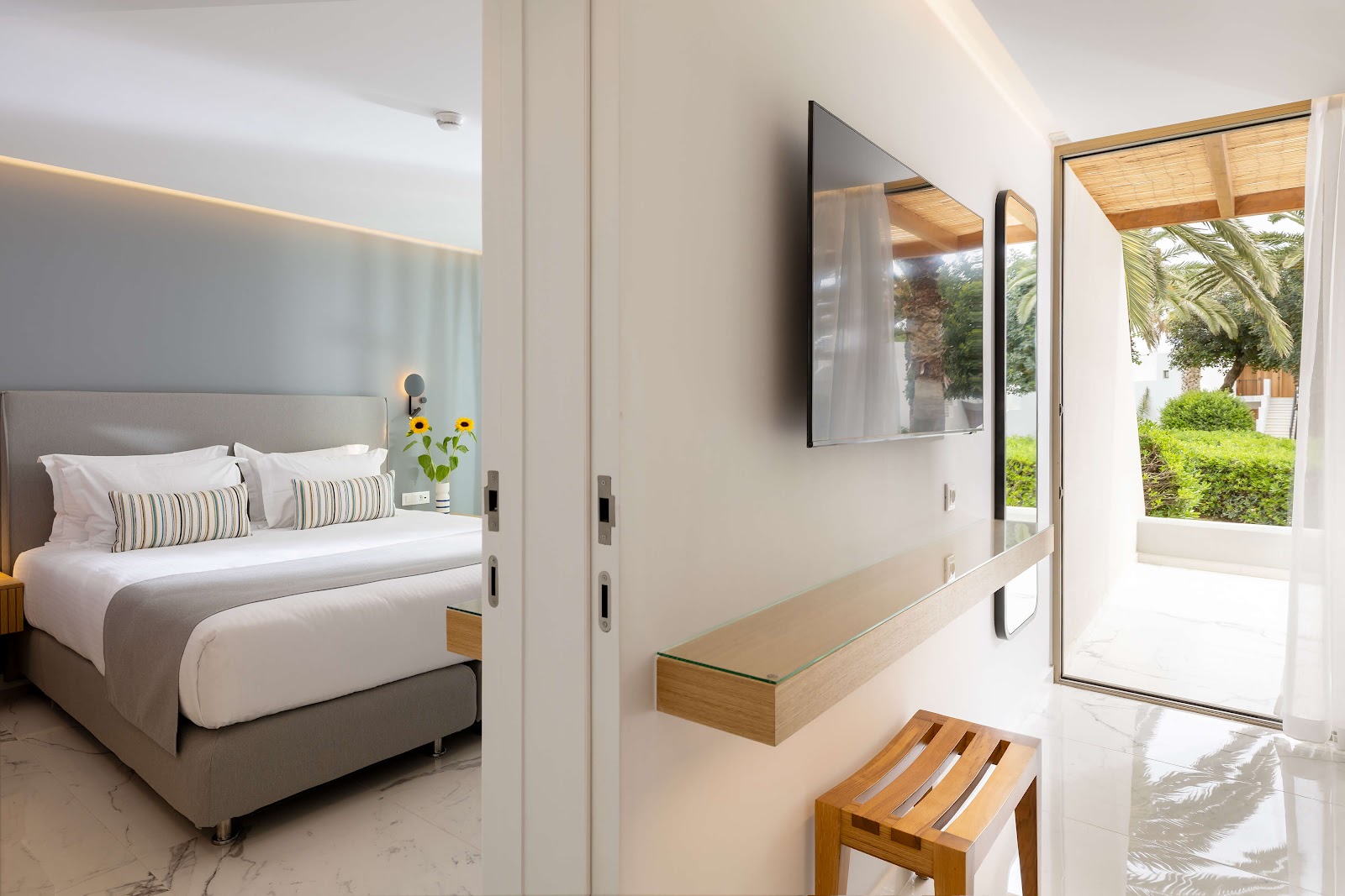 Modern resort bedroom with king bed, wall-mounted TV, and glass doors opening to a patio with palm trees and green grounds.