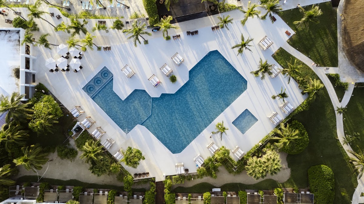 Overhead view of an H-shaped pool with white deck, lounge chairs, and palm trees scattered throughout the resort grounds.