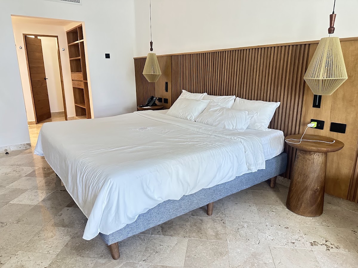 Modern resort bedroom with queen bed, white linens, woven pendant lights, wood nightstand, and open doorway to bathroom.