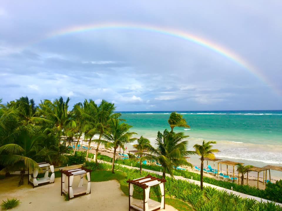 Rainbow arcs over Akumal Bay Beach & Wellness Resort's sandy beach dotted with lounge chairs and palm trees.