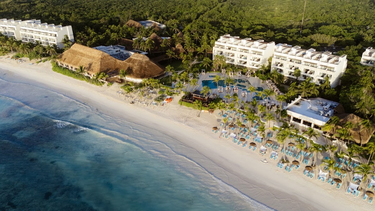 Aerial view of white resort buildings, thatched-roof structures, pools, and blue lounge chairs lining a sandy beach beside turquoise water and tropical forest.