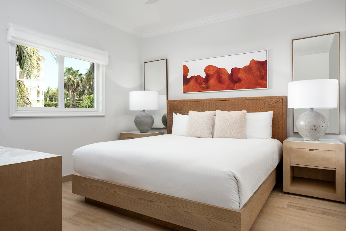 Bright resort bedroom with white walls, wood-frame bed, bedside lamps, and window overlooking palm trees and resort grounds.