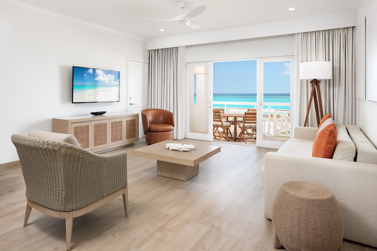 Light-filled resort suite with woven chairs, cream sofa, and ocean view through glass doors opening to a beachfront balcony at Alexandra Resort.
