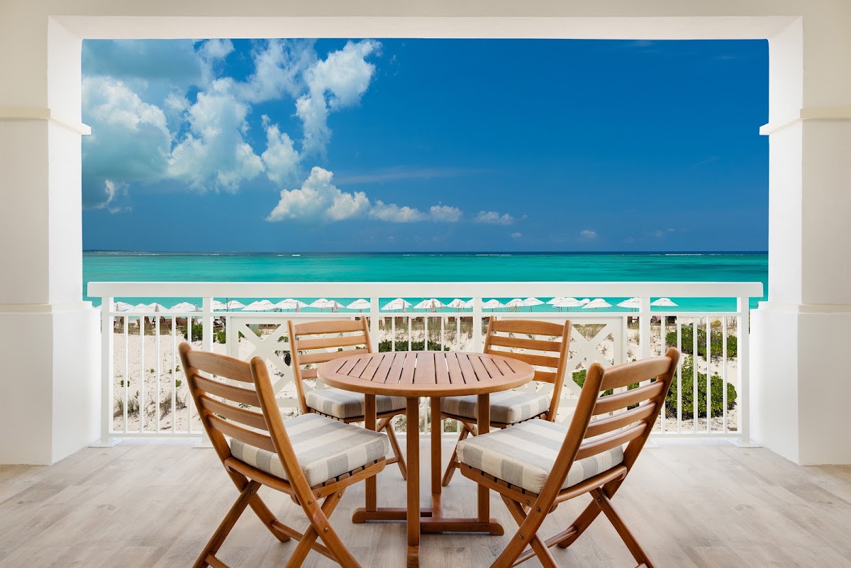 Wooden chairs and round table on a beachfront balcony at Alexandra Resort overlooking turquoise water and white beach umbrellas.