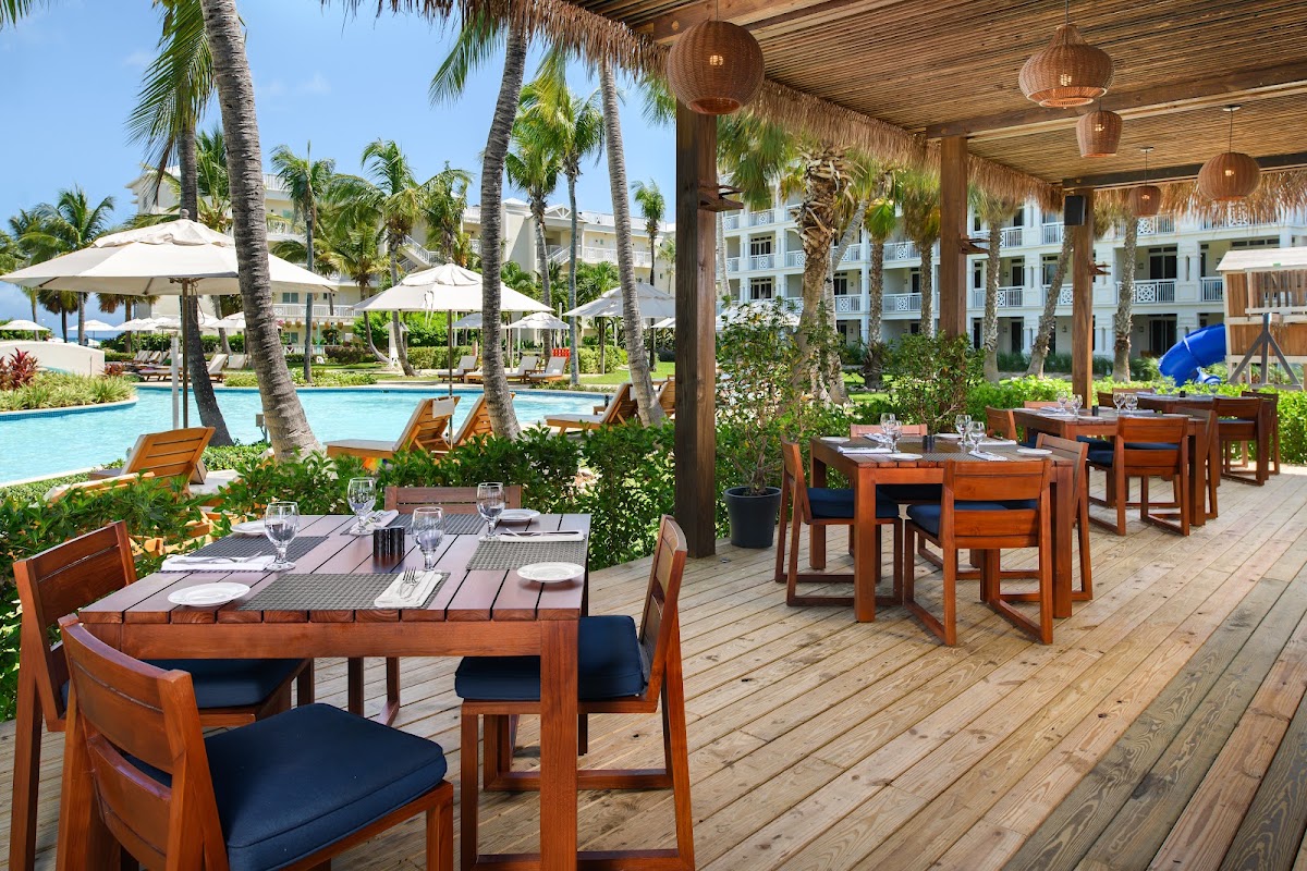 Outdoor dining terrace with wooden tables and chairs overlooking a turquoise pool, palm trees, and white resort buildings under a thatched roof at Alexandra Resort.