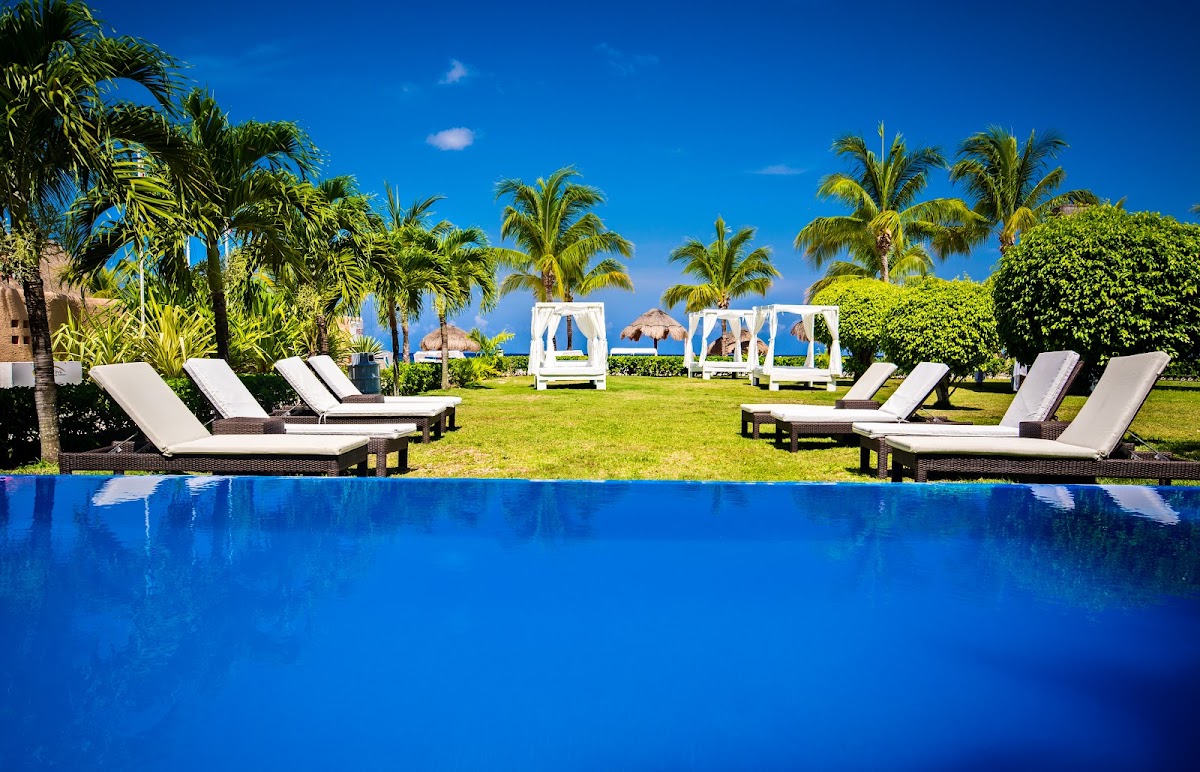 Blue infinity pool with lounge chairs on the lawn, palm trees, white cabanas, and ocean visible beyond at Allegro Cozumel.