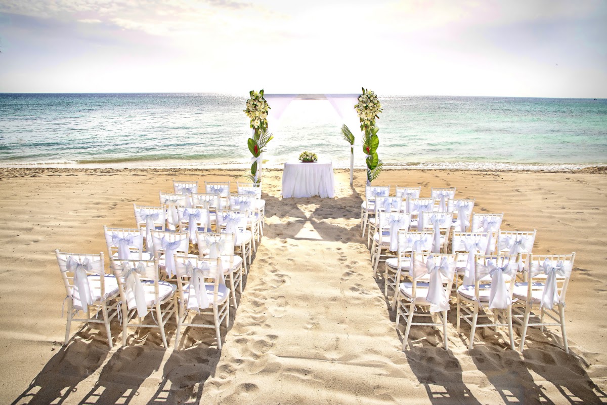 Beach wedding setup with white chairs lining a sandy aisle, floral arch, and ocean horizon at Allegro Cozumel.