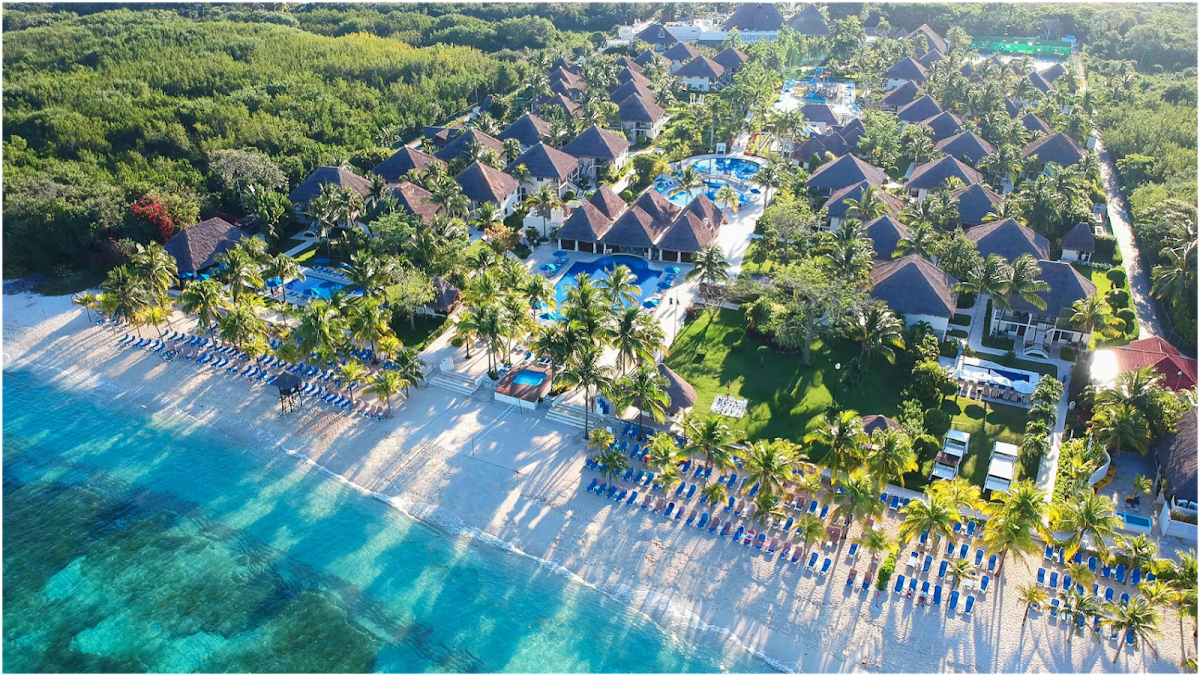 Aerial view of Allegro Cozumel showing turquoise waters, sandy beach with blue loungers, palm trees, multiple thatched-roof bungalows, and swimming pools surrounded by lush vegetation.