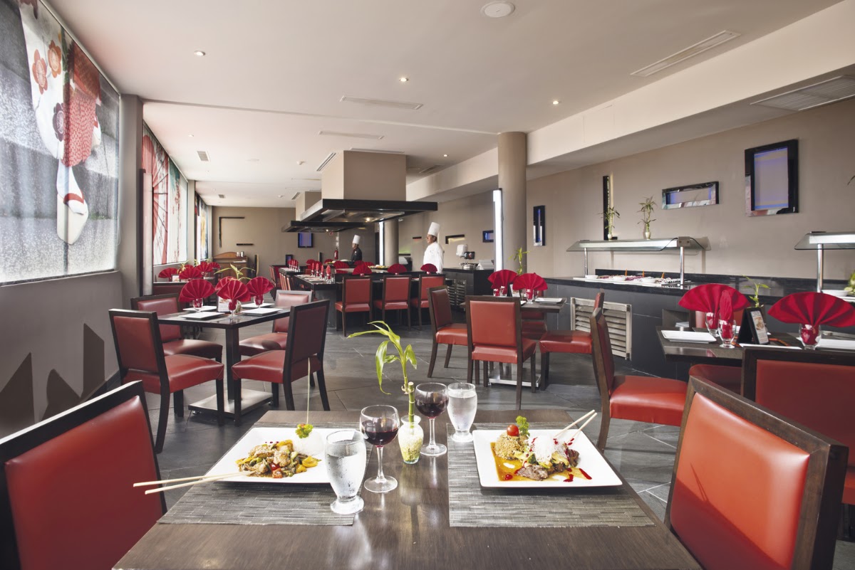 Modern dining room with red leather chairs, black tables set with plated dishes and glasses, red flower centerpieces, and an open kitchen with a chef visible in the background.