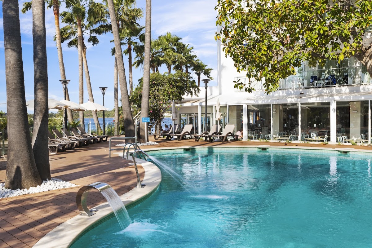 Waterfall feature in turquoise pool surrounded by tall palms, lounge chairs, and white resort building at Alua Boccaccio.