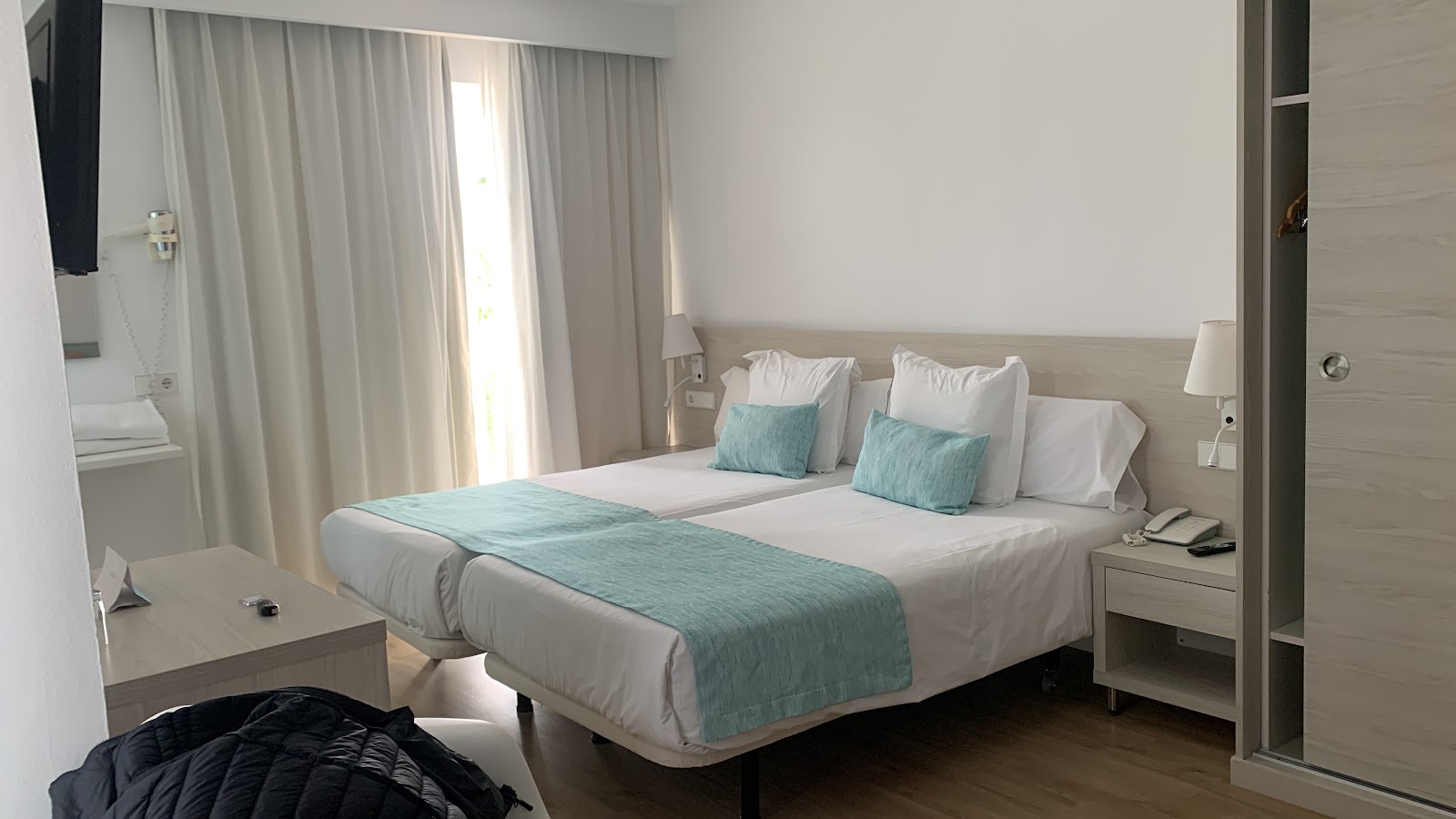 Bright hotel bedroom with two twin beds dressed in white linens and turquoise runner, matching throw pillows, beige headboard, and sheer curtains filtering natural light.