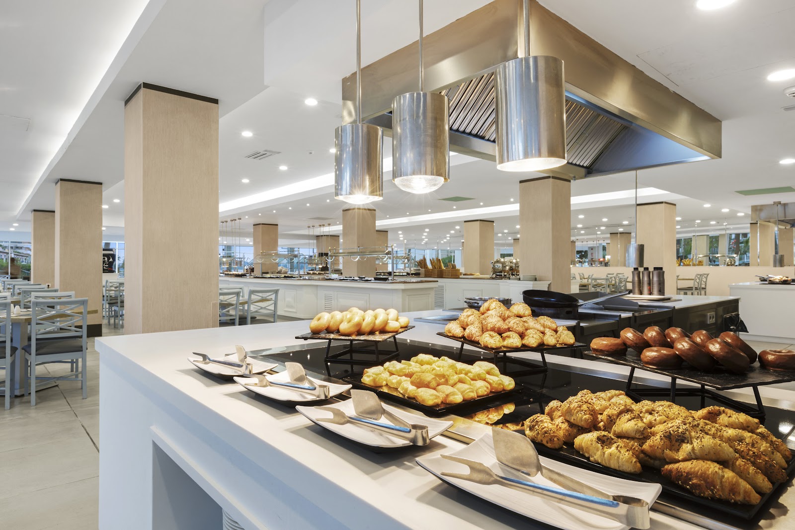 Modern resort buffet with fresh breads, pastries, and rolls displayed on black trays, white counter with place settings, and light wood pillars throughout the dining space.