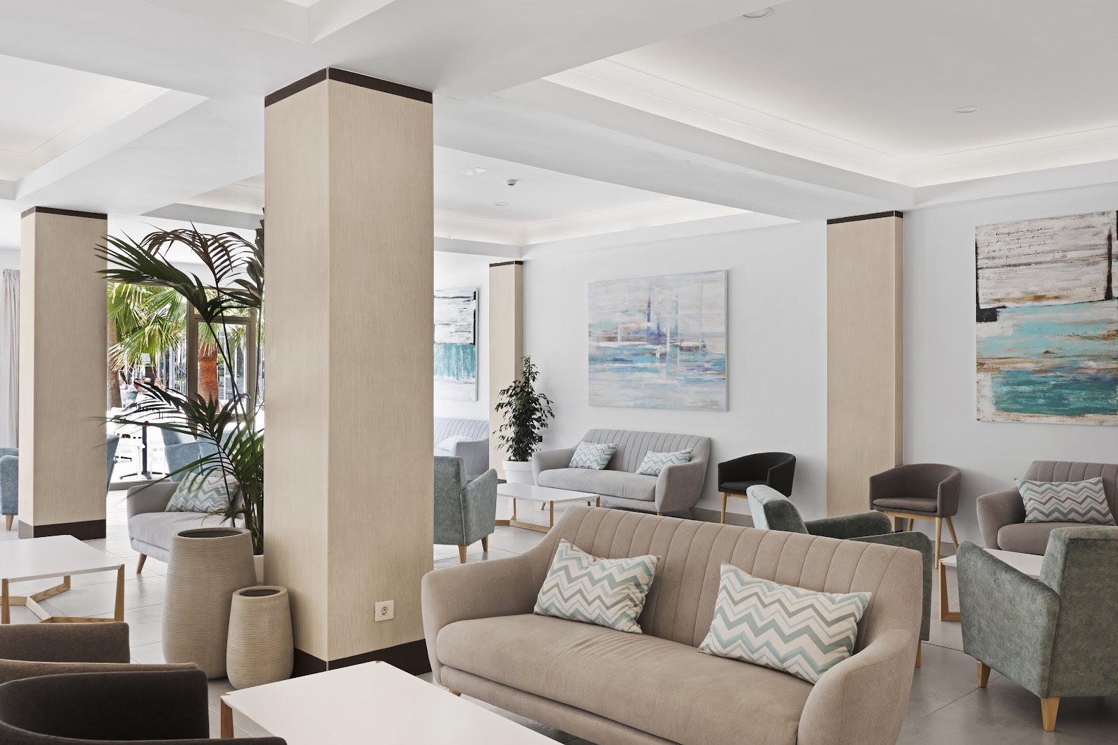 Modern lounge area with beige sofas, tall wooden columns, coastal artwork, and palm plants near floor-to-ceiling windows.