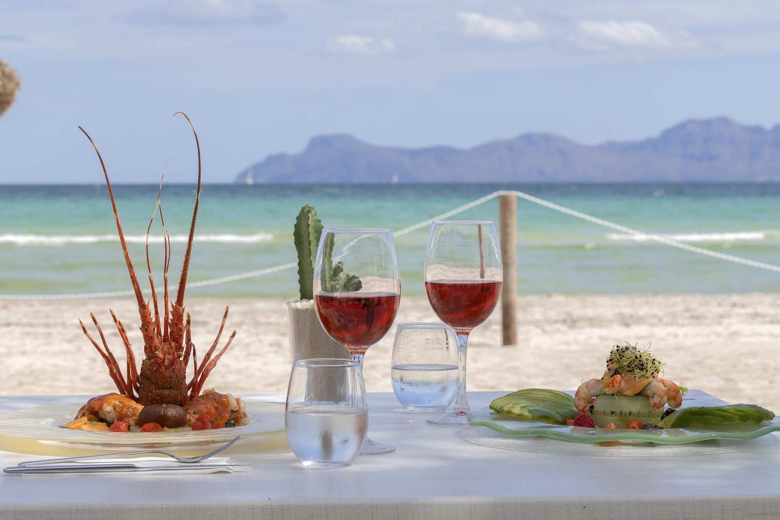 Beachfront dining table at Alua Boccaccio with red wine, seafood appetizers, and ocean view with mountains beyond.