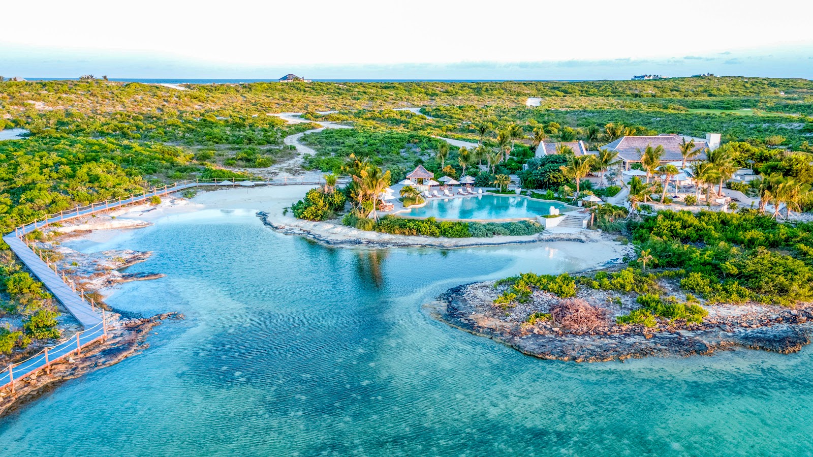 Aerial view of Ambergris Cay showing turquoise lagoon, resort pool surrounded by palm trees, thatched-roof buildings, and lush mangrove forest beyond.