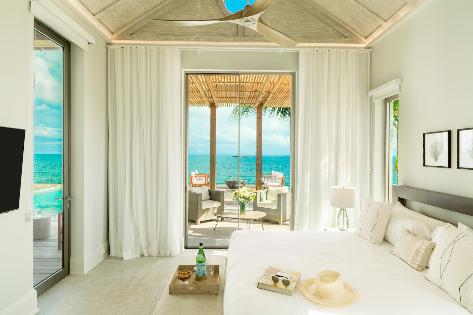 Bright beachfront villa bedroom with white curtains and wooden ceiling, opening onto a furnished terrace overlooking turquoise ocean water.