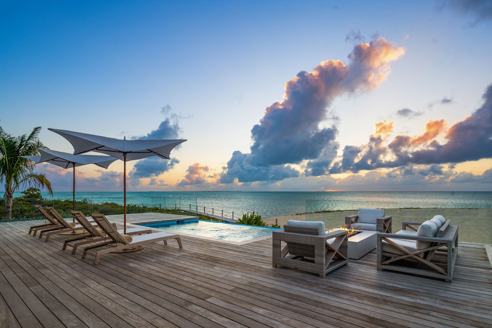 Beachfront villas with thatched roofs sit on a lush, vegetated island overlooking turquoise waters under a partly cloudy sky.
