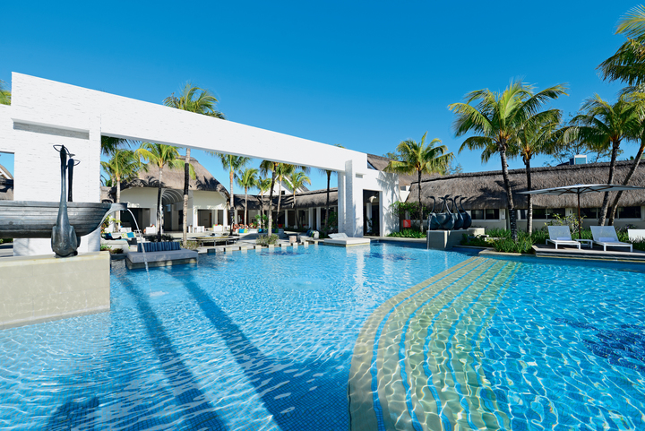 Large resort pool with clear blue water, white modern buildings, palm trees, and thatched-roof structures under a clear sky.
