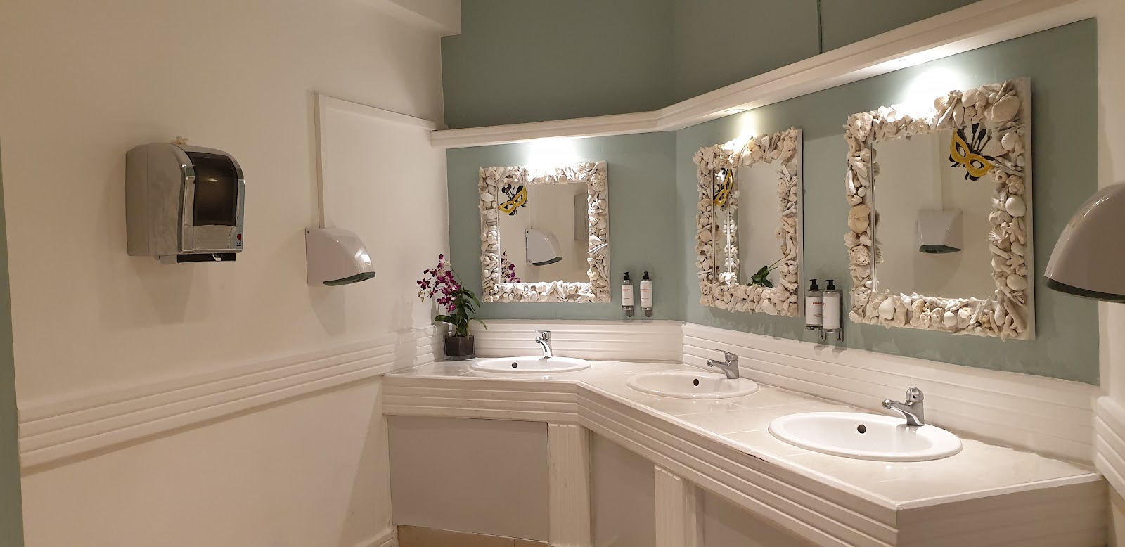 Resort bathroom with three sinks, ornate framed mirrors, sage green walls, and white wainscoting at Ambre, A Sunlife Resort.