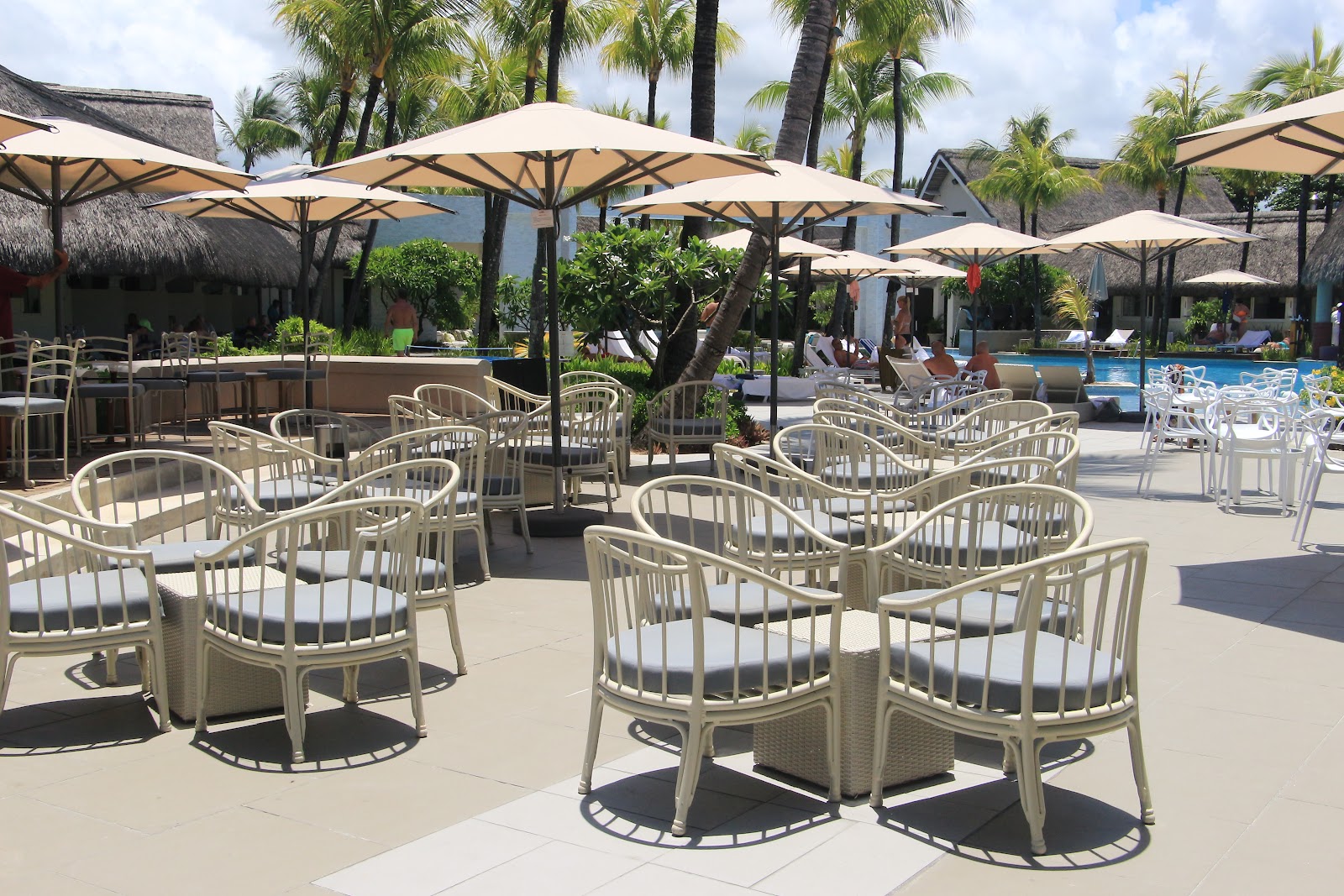 Beige woven chairs with cushions arranged on a patio under cream umbrellas, with palm trees, thatched-roof bungalows, and a pool visible beyond.