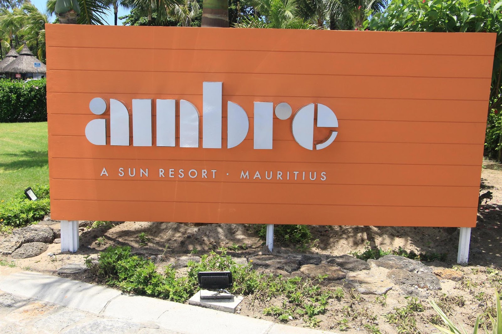 Orange sign with white lettering reading "ambre A SUN RESORT MAURITIUS" surrounded by tropical palms and landscaping.