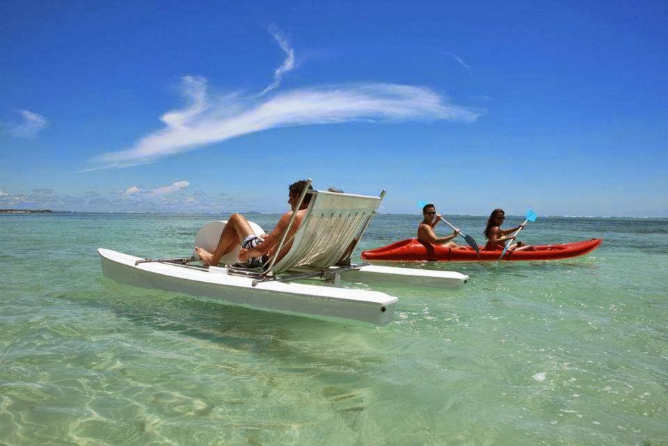 A man reclines in a deck chair on an outrigger canoe while two women paddle a red kayak nearby in shallow turquoise water.