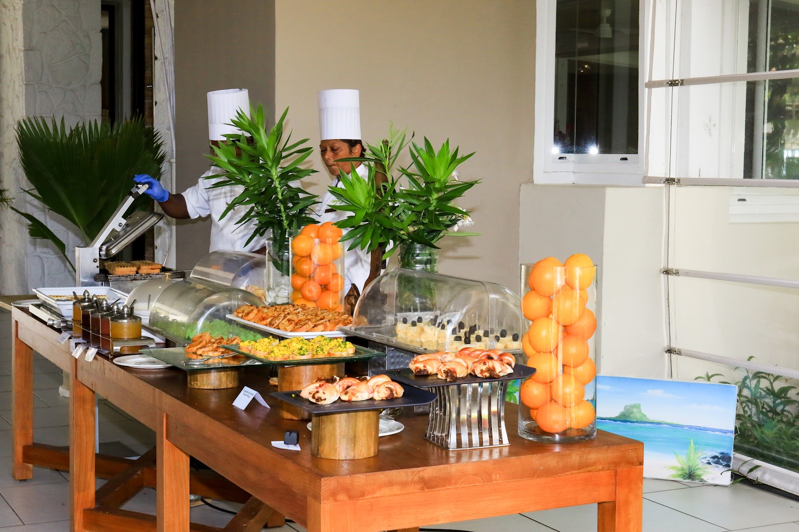 Chef in tall white hat stands behind wooden buffet table displaying pastries, fresh oranges in glass vases, and tropical plants on a beige building patio.