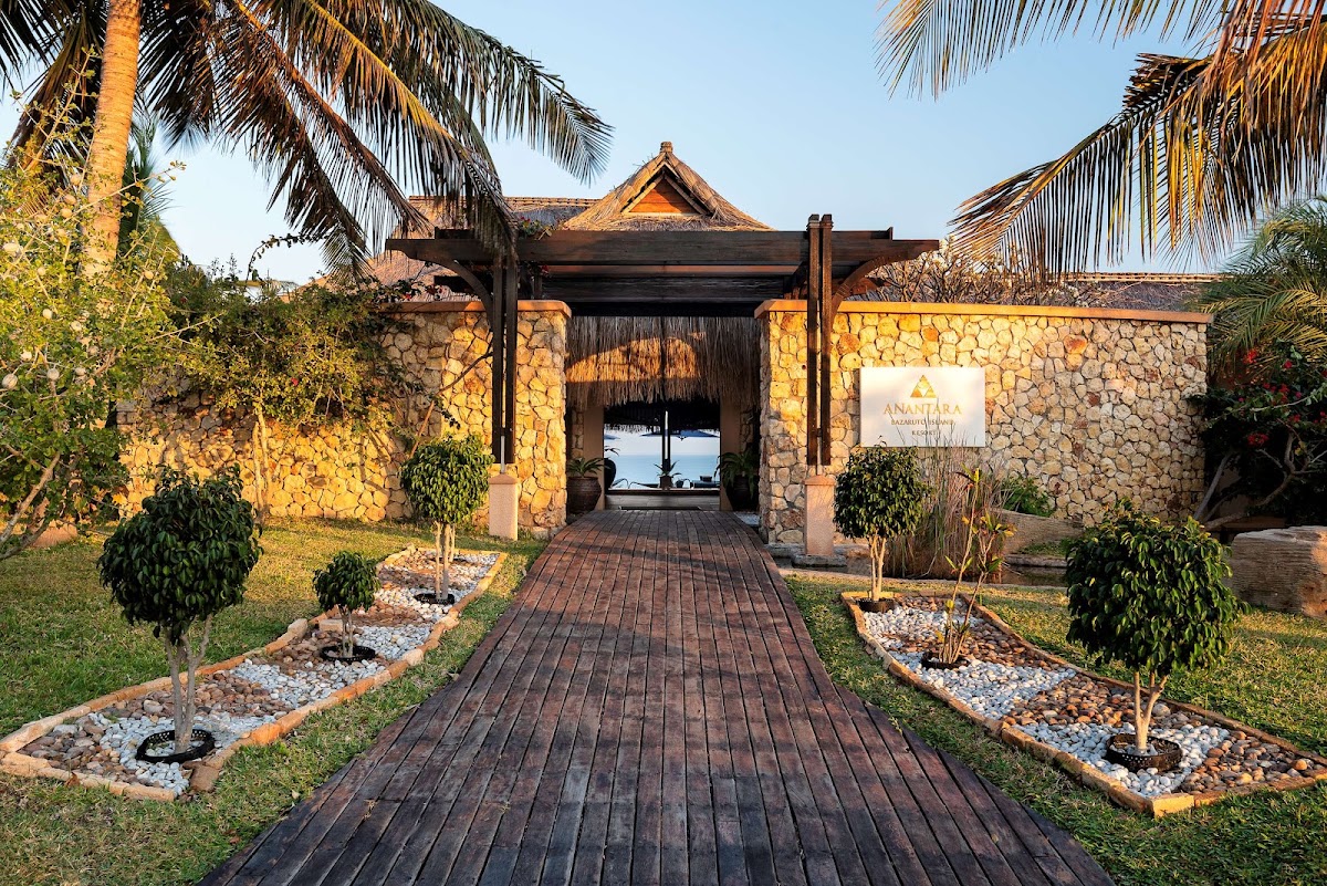 Wooden boardwalk leads through palm-lined grounds to stone entrance gateway of Anantara Bazaruto Island Resort, with ocean visible beyond.