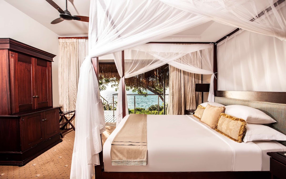 Beachfront bedroom with four-poster canopy bed draped in white sheer fabric, ocean view through open doors, dark wood furniture, and ceiling fan.