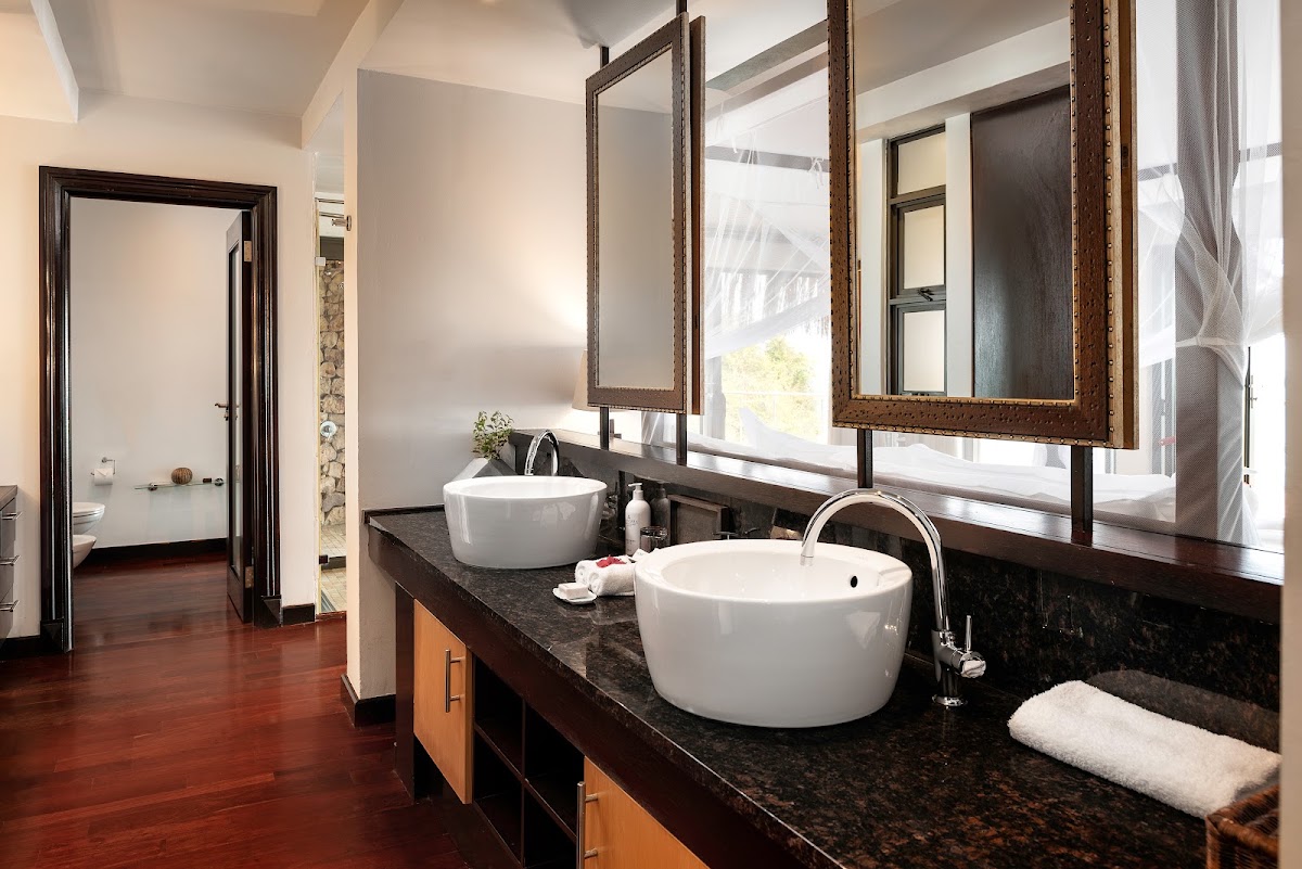 Resort bathroom with dual white round sinks, dark granite countertop, framed mirrors, and dark wood flooring leading to separate toilet room.