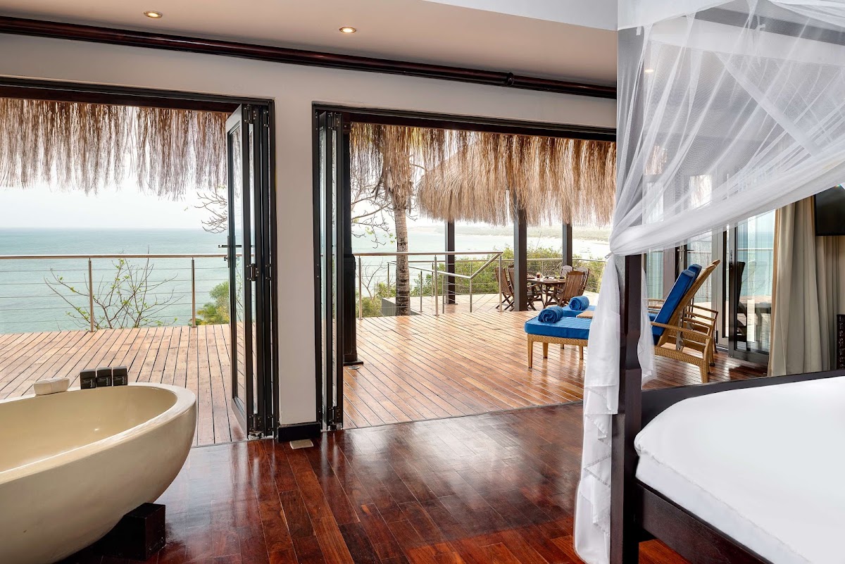 Beachfront resort suite with wooden floors, freestanding bathtub, and open doors to a teak deck overlooking the Indian Ocean and thatched-roof structures.