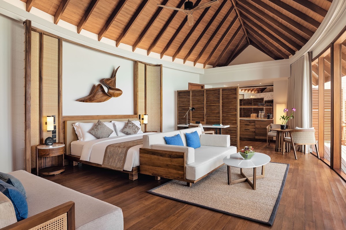 Spacious resort villa interior with wooden beam ceiling, king bed with blue accent pillows, lounge seating, wooden desk, and tropical sculpture on white wall.