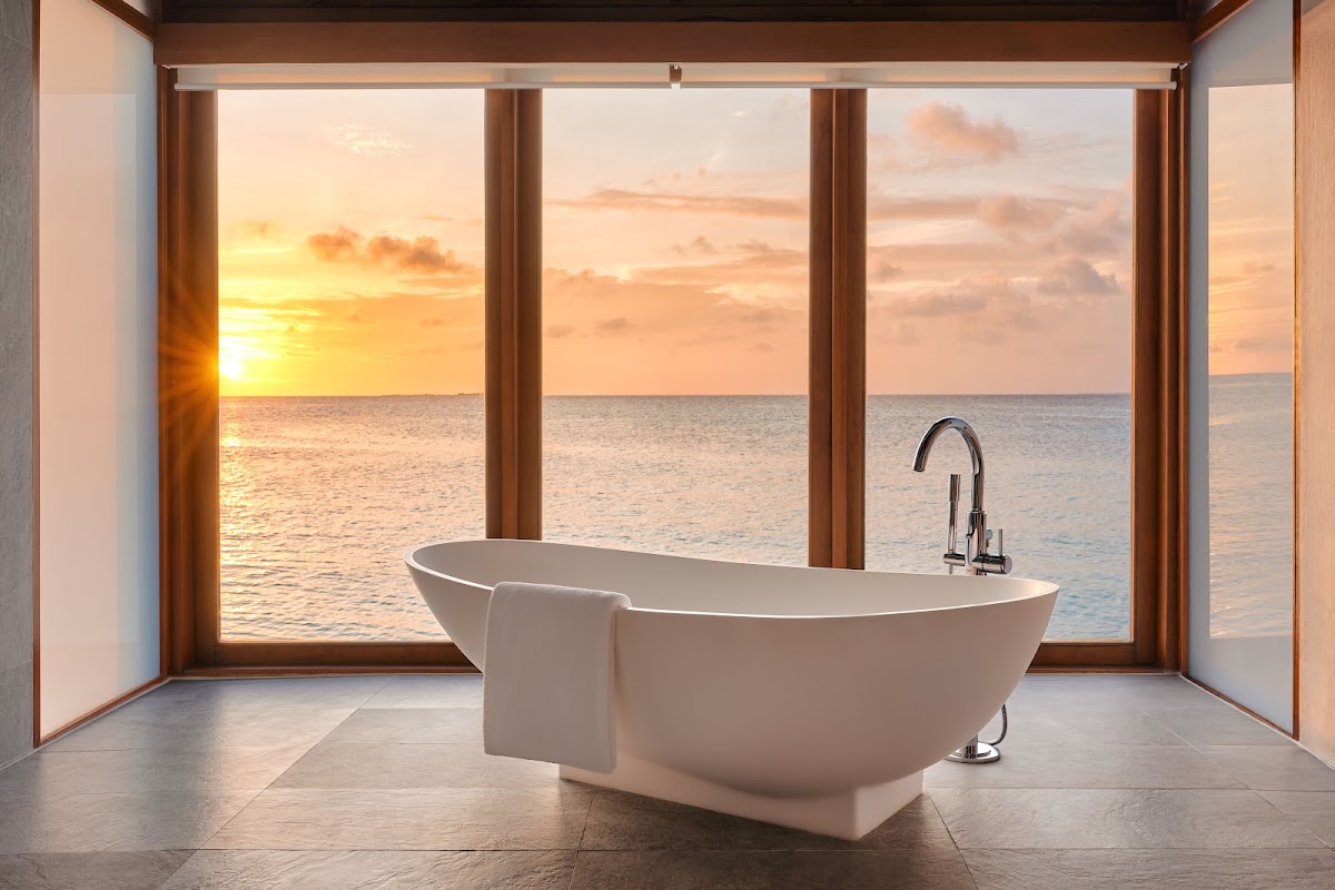 White freestanding bathtub with chrome faucet in a bathroom with large windows framing an ocean sunset at Anantara Dhigu Maldives Resort.