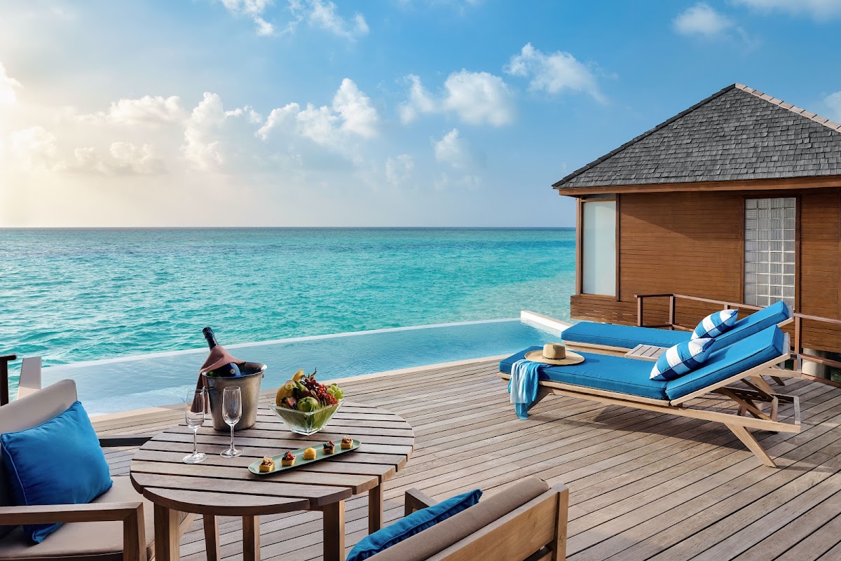 Overwater bungalow with wooden deck, blue loungers, dining table with wine and fruit, and turquoise ocean under clear sky.