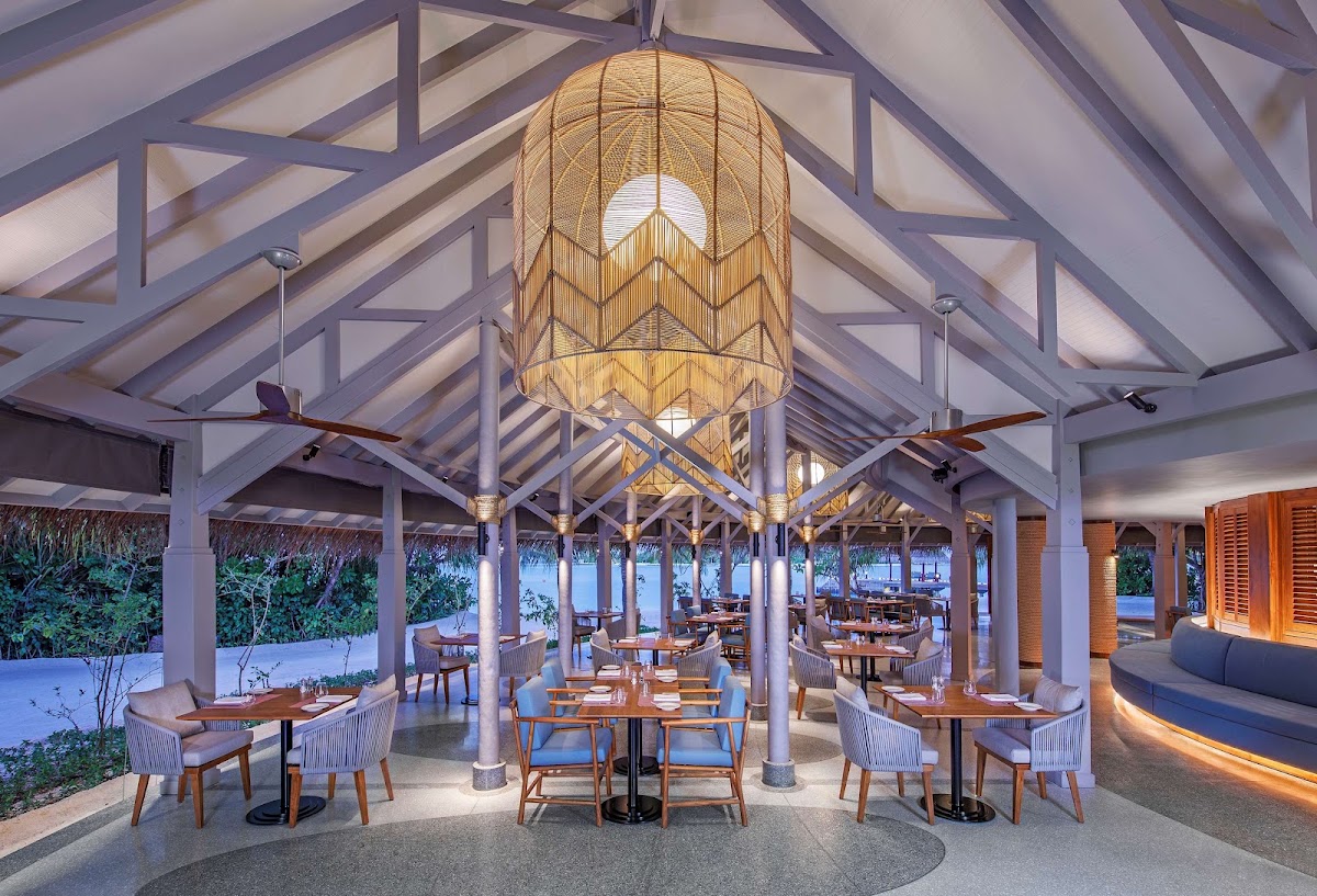 Open-air dining pavilion with woven pendant lamp, wooden tables, upholstered chairs, and ocean views through supporting columns at Anantara Dhigu Maldives Resort.