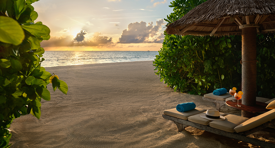 Thatched cabana with loungers and towels overlooks a sandy beach and ocean at sunset, framed by tropical foliage.
