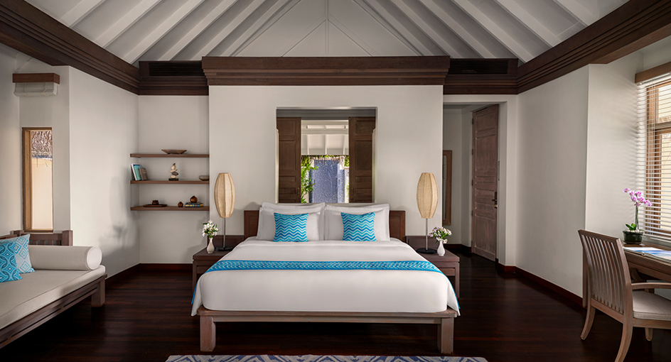 Spacious resort bedroom with dark wood bed frame, white linens, turquoise pillows, vaulted ceiling with exposed beams, and ocean view through central doorway.