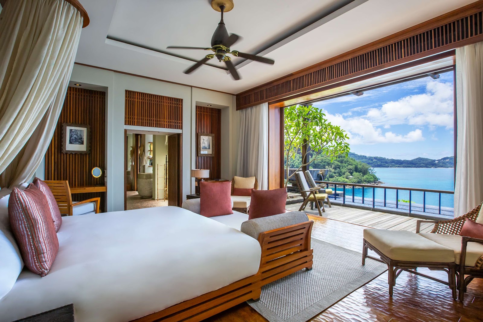 Spacious resort bedroom with wooden furnishings, white bedding, and floor-to-ceiling windows overlooking a turquoise bay and forested coastline.