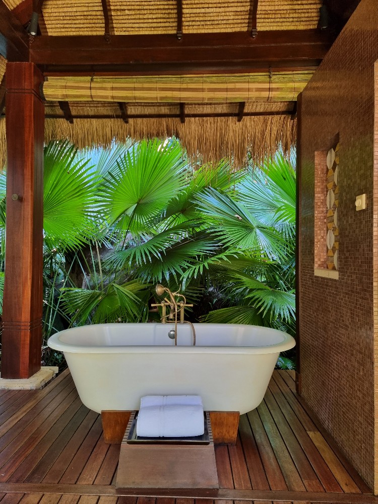 Freestanding bathtub on wooden deck inside open-air pavilion with thatched roof, surrounded by lush green palm fronds and brick walls.