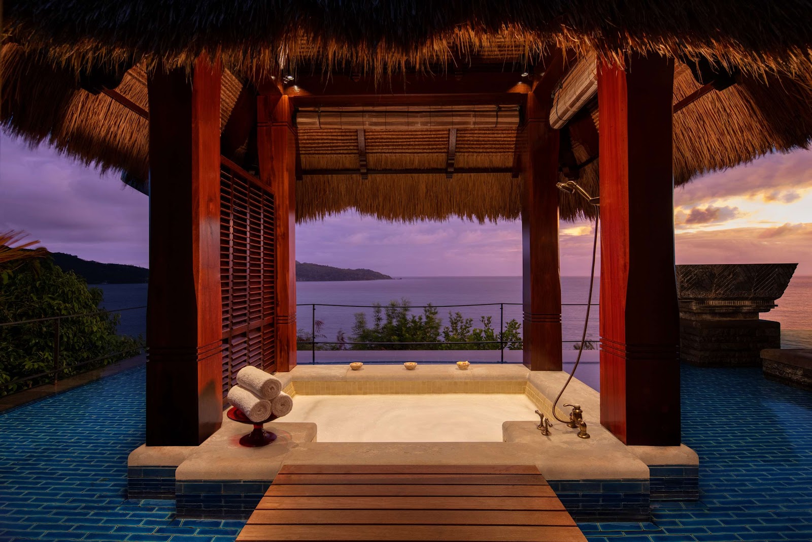 Spa therapist performs a massage on a guest at an outdoor treatment pavilion surrounded by tropical palms and a large rock formation.