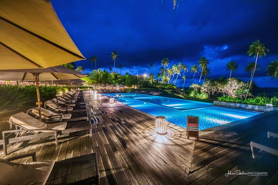 Illuminated pool deck at night with wooden loungers under yellow umbrellas, palm trees, and ocean view at Anantara Peace Haven Tangalle Resort.