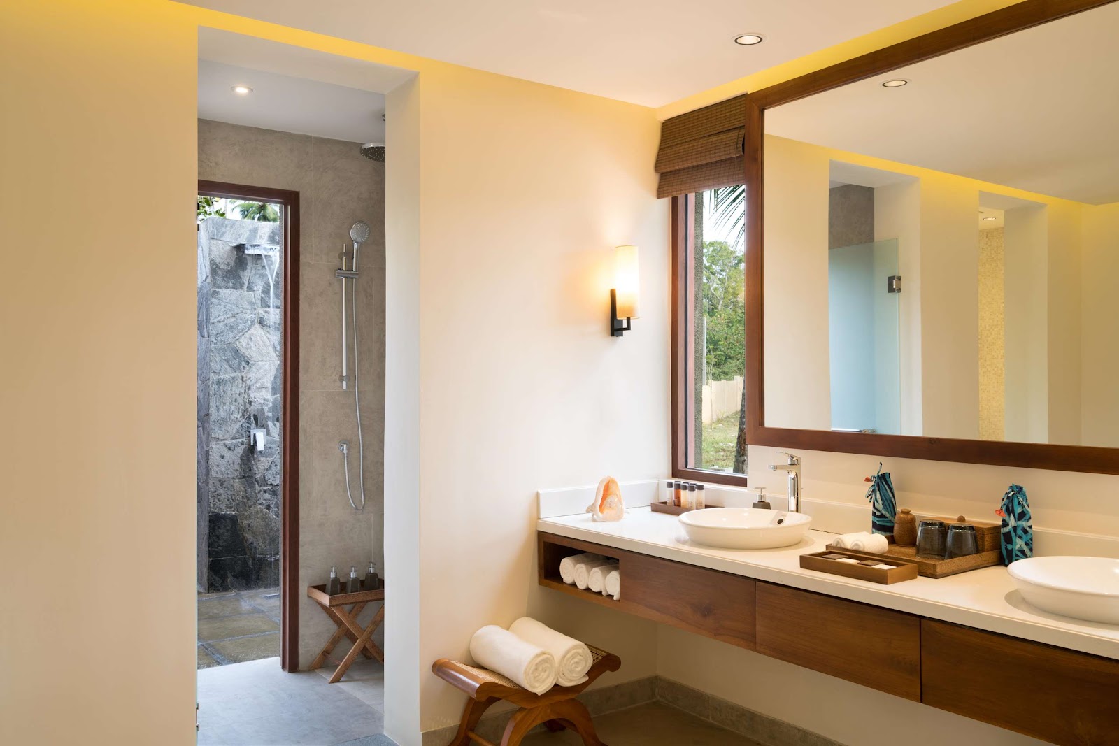 Resort bathroom with cream walls, wooden vanity, round white sink, large mirror, and stone shower enclosure visible through doorway.