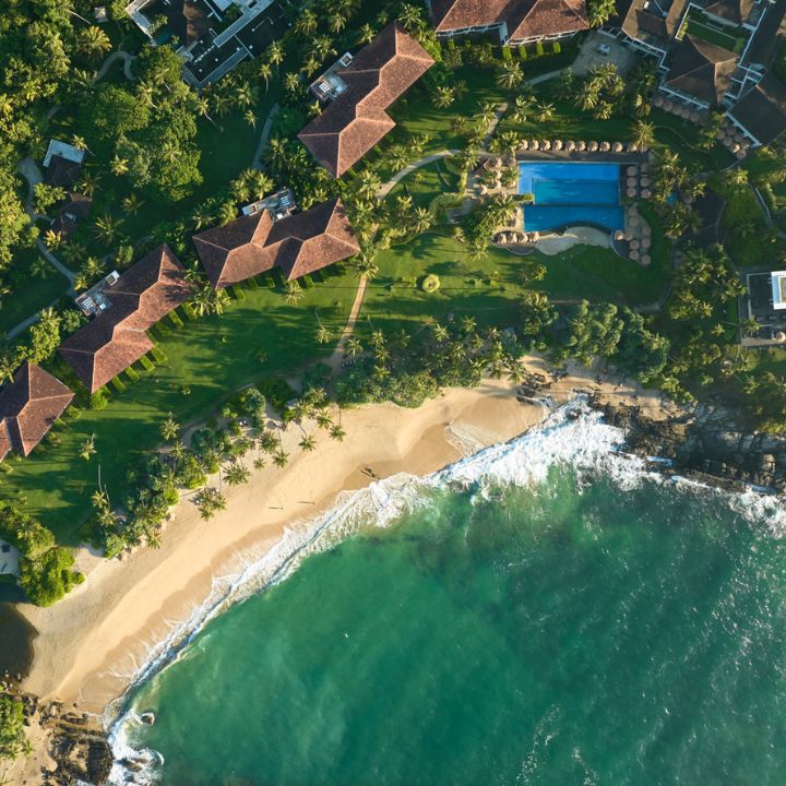 Aerial view of Anantara Peace Haven Tangalle Resort with tiled-roof bungalows, manicured grounds, blue pool, and sandy beach meeting turquoise ocean waves.