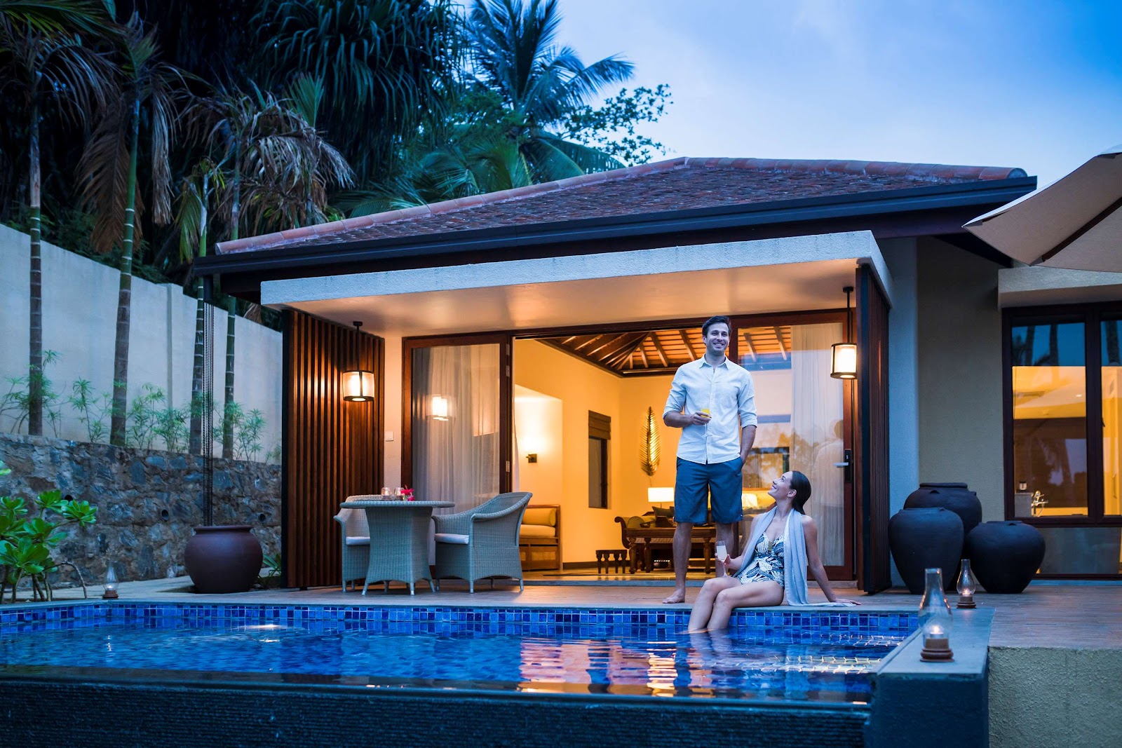 Woman sits at edge of illuminated blue pool while man stands on terrace of modern villa at dusk, with palm trees and stone walls visible.