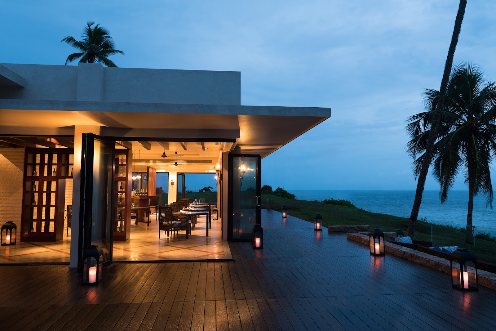 Modern oceanfront dining pavilion at dusk with lit lanterns, wooden deck, and ocean views beyond palm trees.
